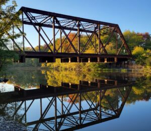 This color photo from 2021 shows the Grand Trunk rail bridge over the calm, reflective Royal River, surrounded by trees with autumn foliage.