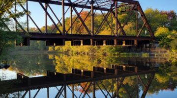 This color photo from 2021 shows the Grand Trunk rail bridge over the calm, reflective Royal River, surrounded by trees with autumn foliage.